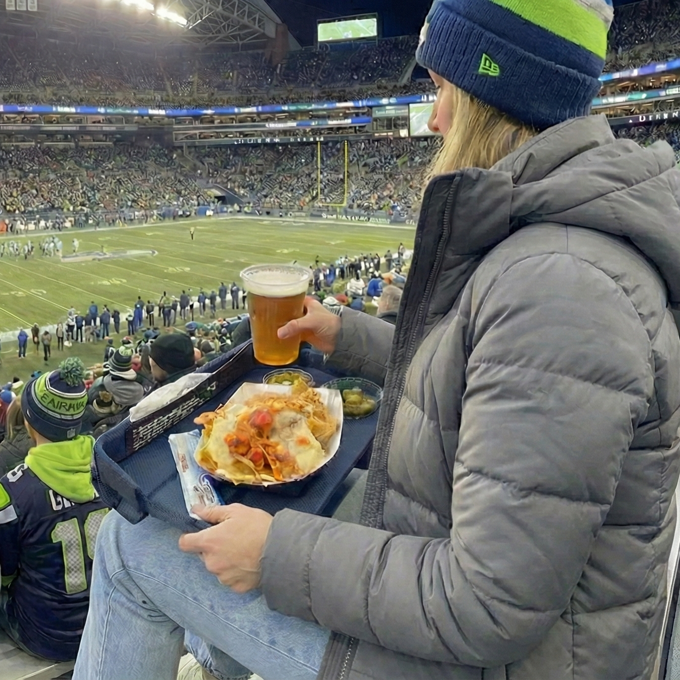 Person watching a football game in a stadium with others, holding a beer and food on a GoTray.