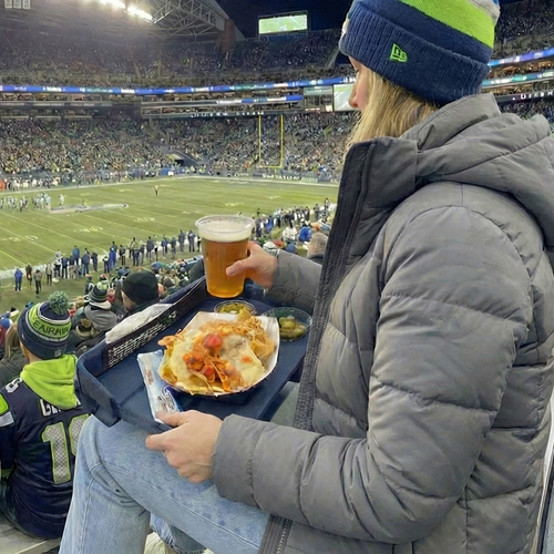 Person watching a football game in a stadium with others, holding a beer and food on a GoTray.